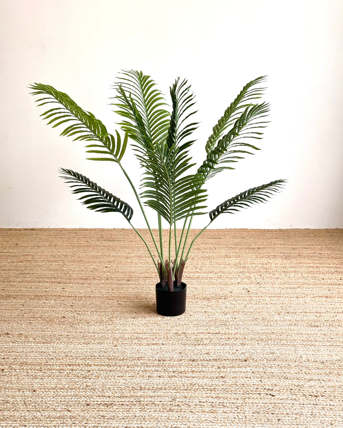 An artificial green palm frond plant in a small black pot, set against a neutral beige woven rug and plain white wall background.