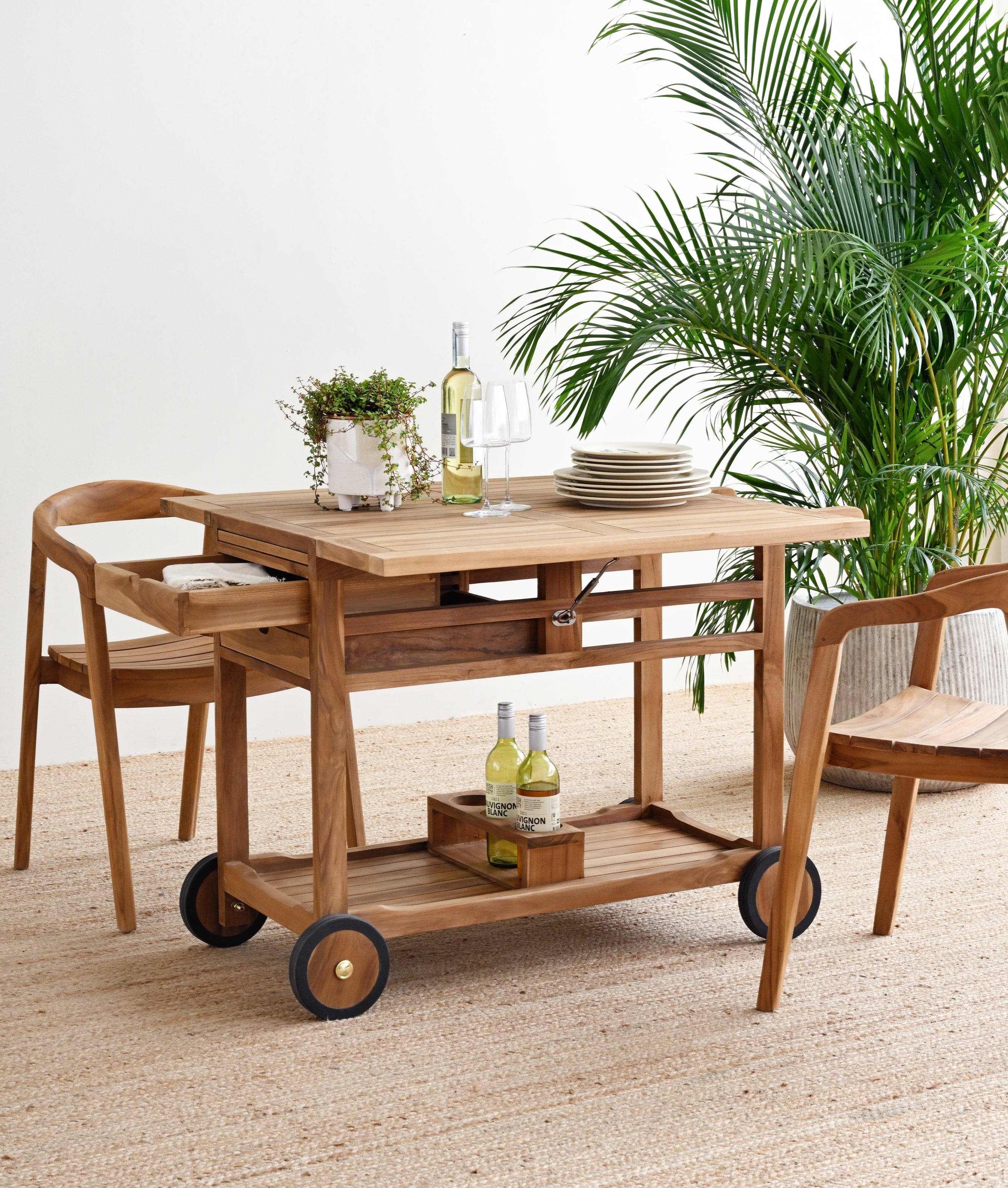 Wooden serving cart with drinks and plates on a beige carpet, surrounded by plants.