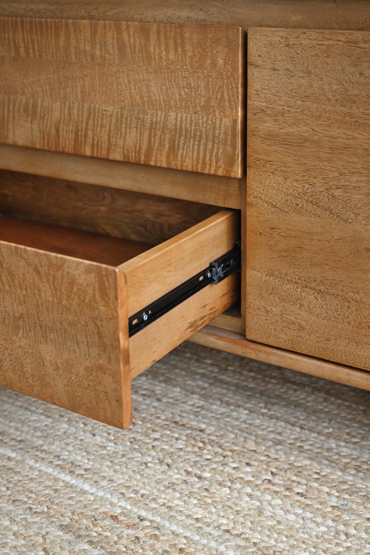 Close-up of a wooden dresser or cabinet with one drawer pulled open, showing black metal drawer hardware on a woven rug.