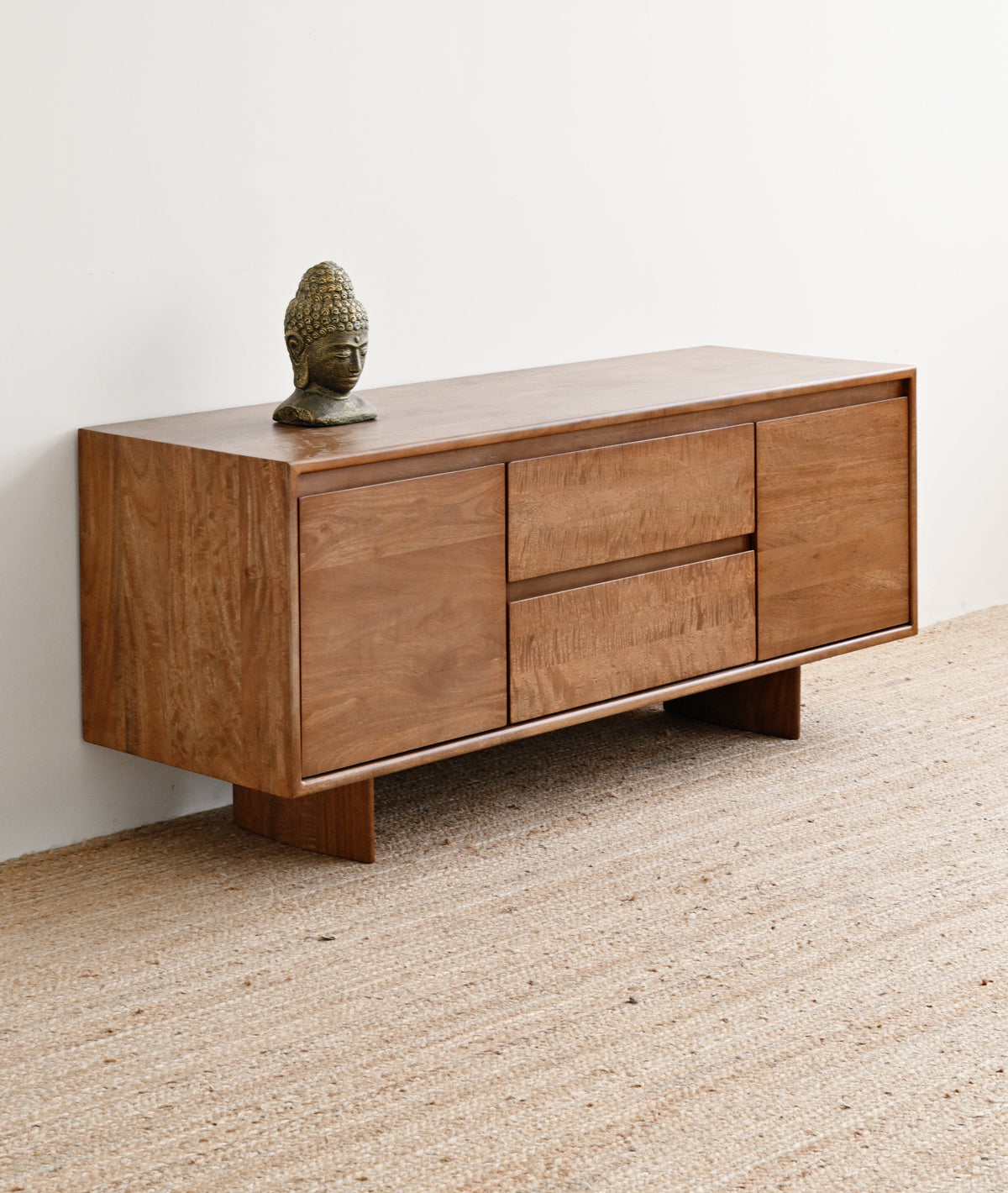 A mid-century modern wooden sideboard with a warm medium-brown finish, featuring two drawers on the left and two cabinet doors on the right, styled on a woven jute rug against a plain white wall.