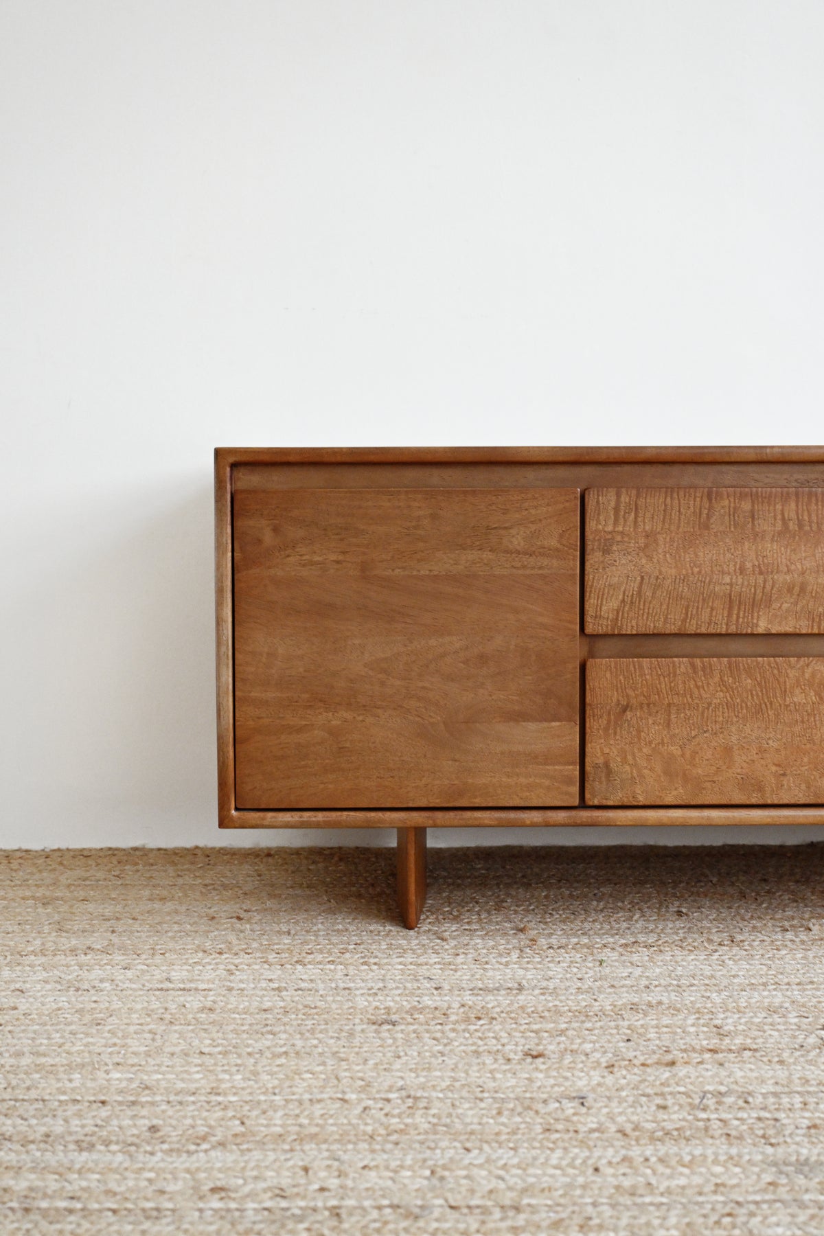 A mid-century modern wooden sideboard with a warm medium-brown finish, featuring a single cabinet door and two drawers on the right, supported by a tapered leg, set against a white wall and a textured beige rug.