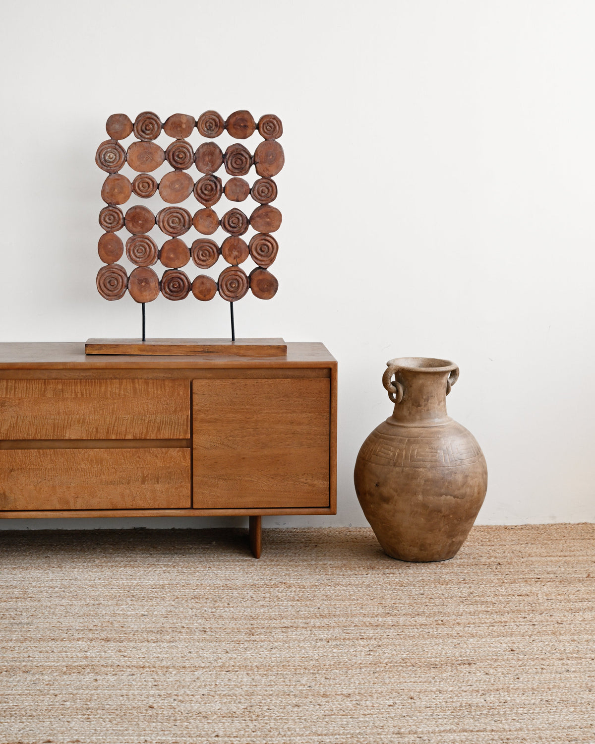 A mid-century modern wooden sideboard cabinet with a decorative wooden wall art piece on top, set against a white wall on a beige woven rug next to a large brown ceramic vase.