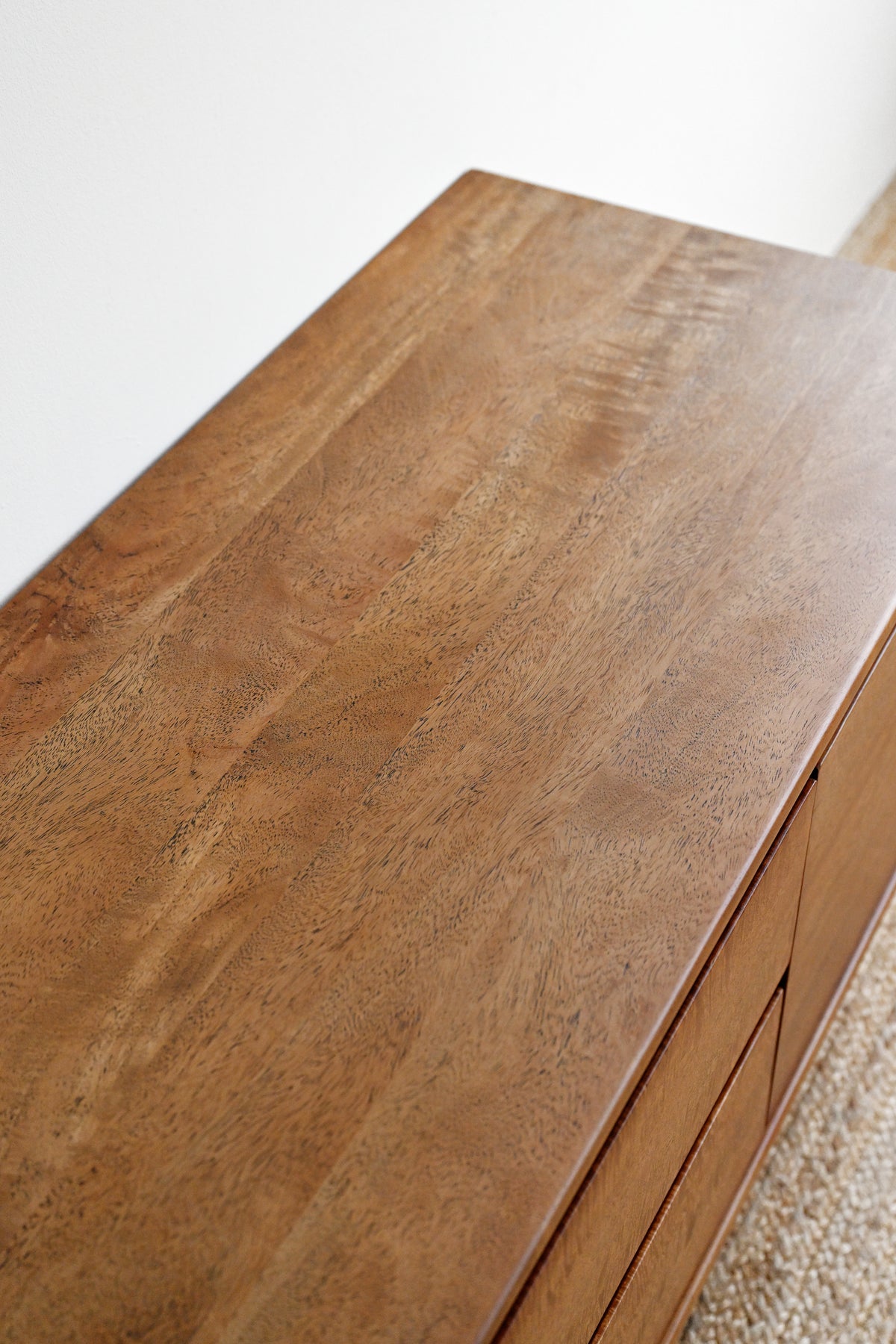 Close-up of a medium brown wooden dresser or cabinet top with visible wood grain, photographed indoors against a light wall and carpet.