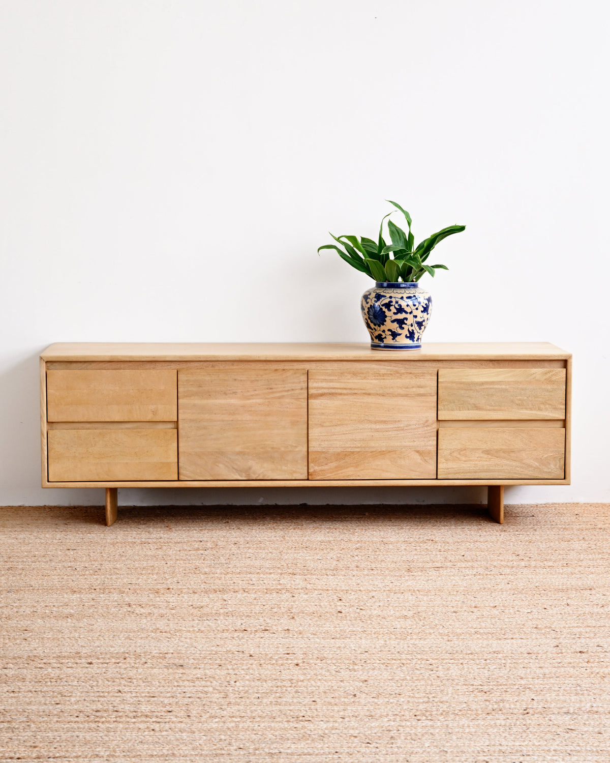 A light natural wood sideboard cabinet with multiple drawers and doors, styled with a small potted green plant on top, set against a plain white wall and a textured beige woven rug.