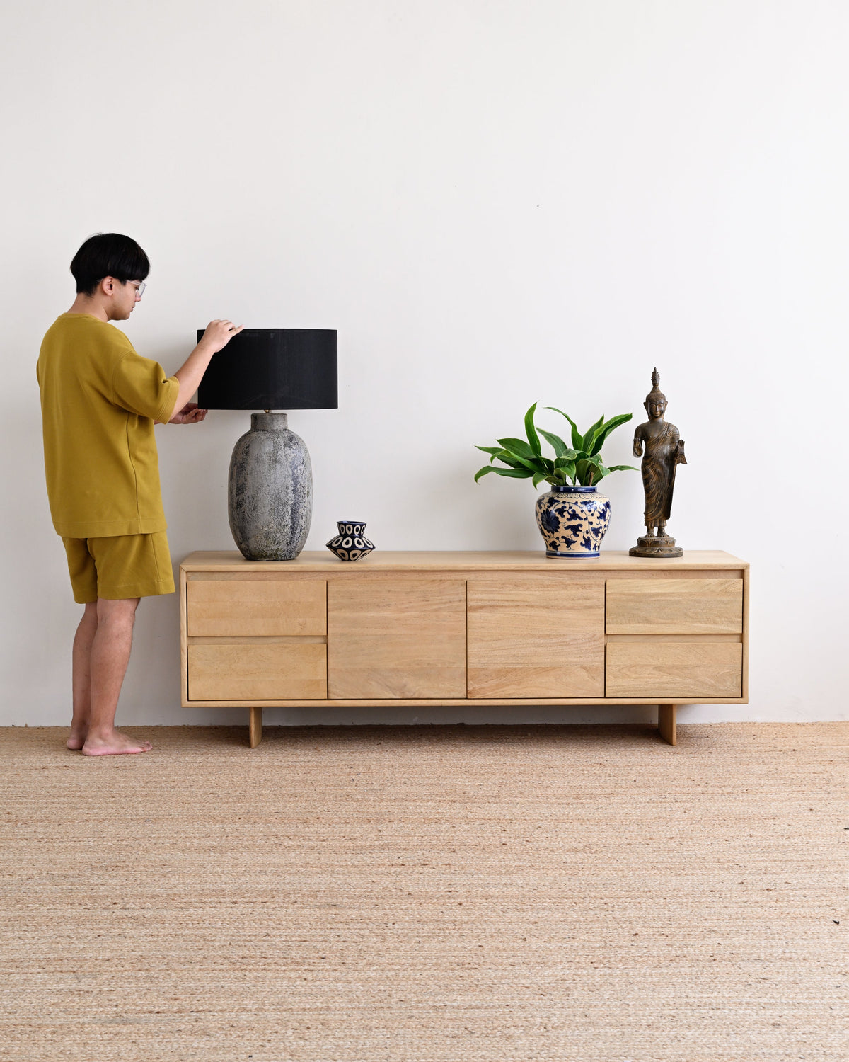 A man in a mustard yellow t-shirt and shorts stands next to a light wood sideboard, adjusting a black lampshade on a gray ceramic lamp. The room has a white wall and a light beige woven rug.