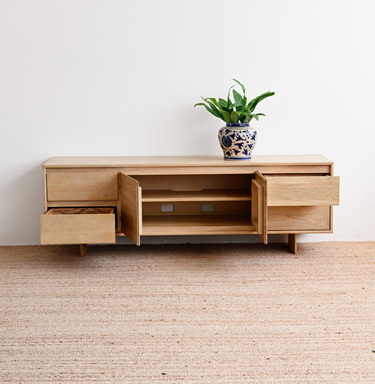 A light natural wood TV stand or console cabinet with open drawers and shelving, placed on a woven beige rug against a plain white wall. A small green potted plant sits on top.
