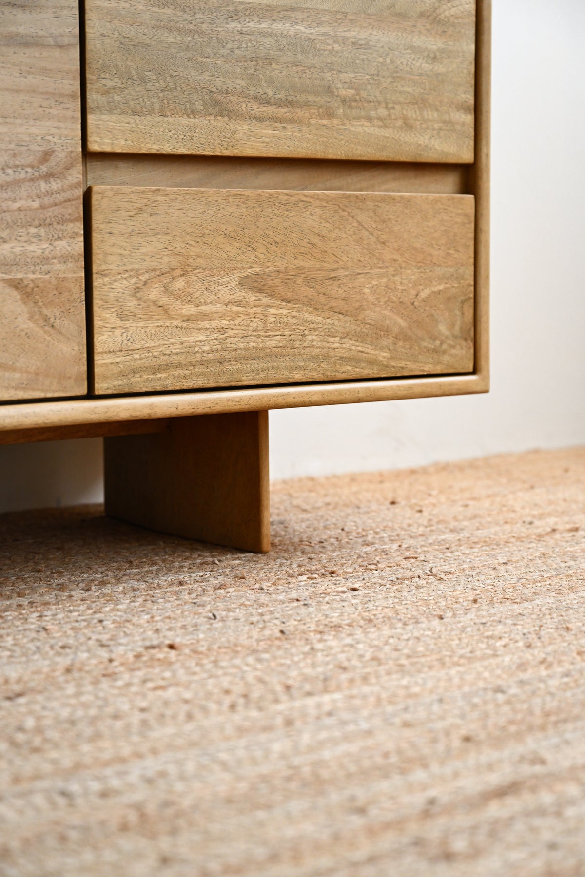 Close-up of a light natural wood furniture piece with a visible grain, showing part of a drawer or cabinet and its support leg above a woven beige rug.