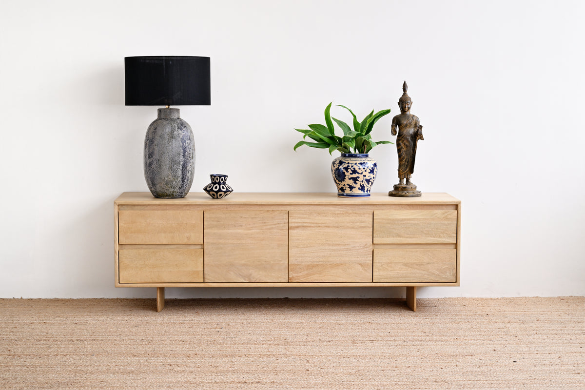 A mid-century modern light oak wood sideboard cabinet styled with a black lamp, a potted green plant, and a bronze Buddha statue against a white wall and beige jute rug.