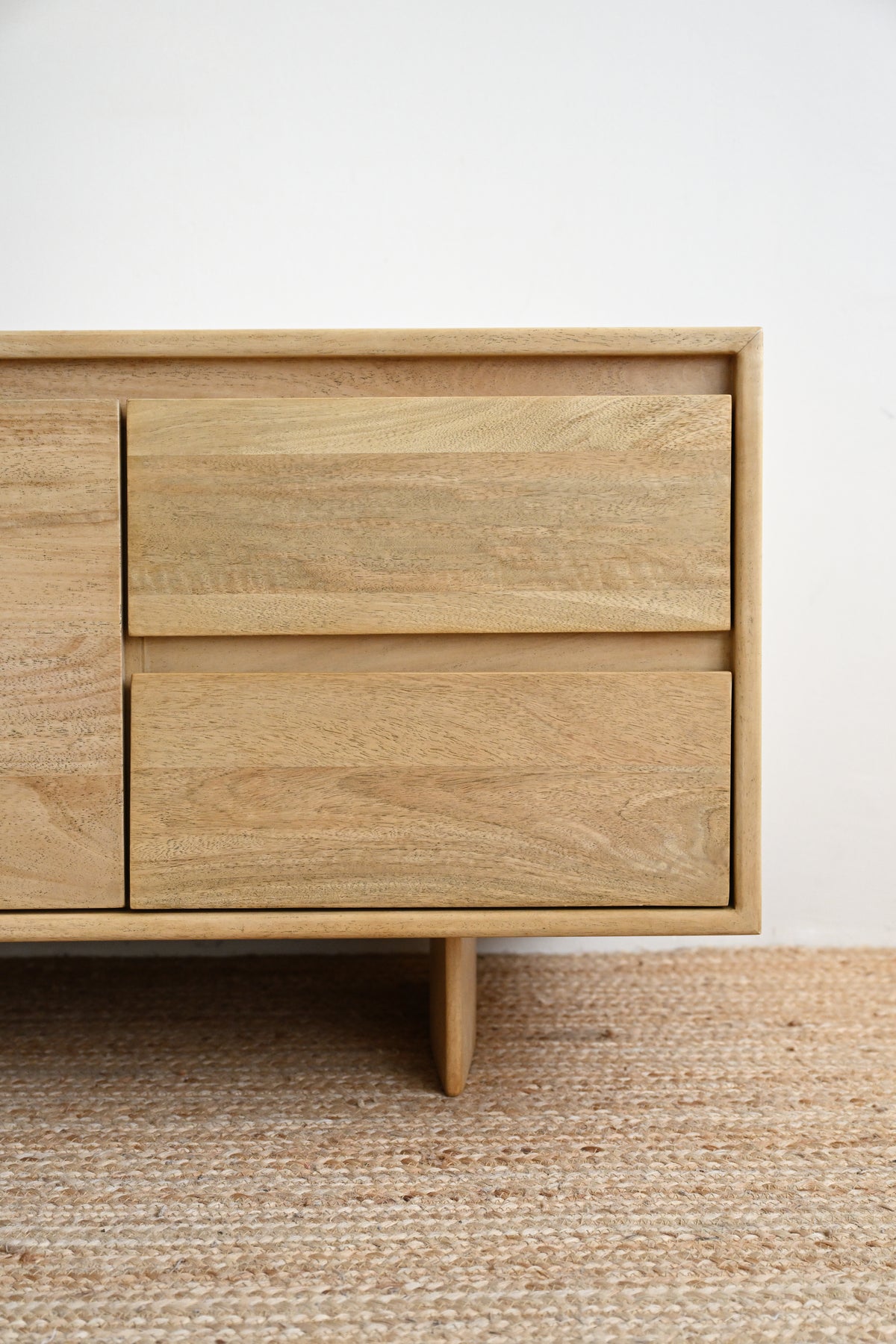 Close-up of a light natural wood sideboard or dresser with two drawers and a tapered leg, set against a plain white wall and a woven beige rug.