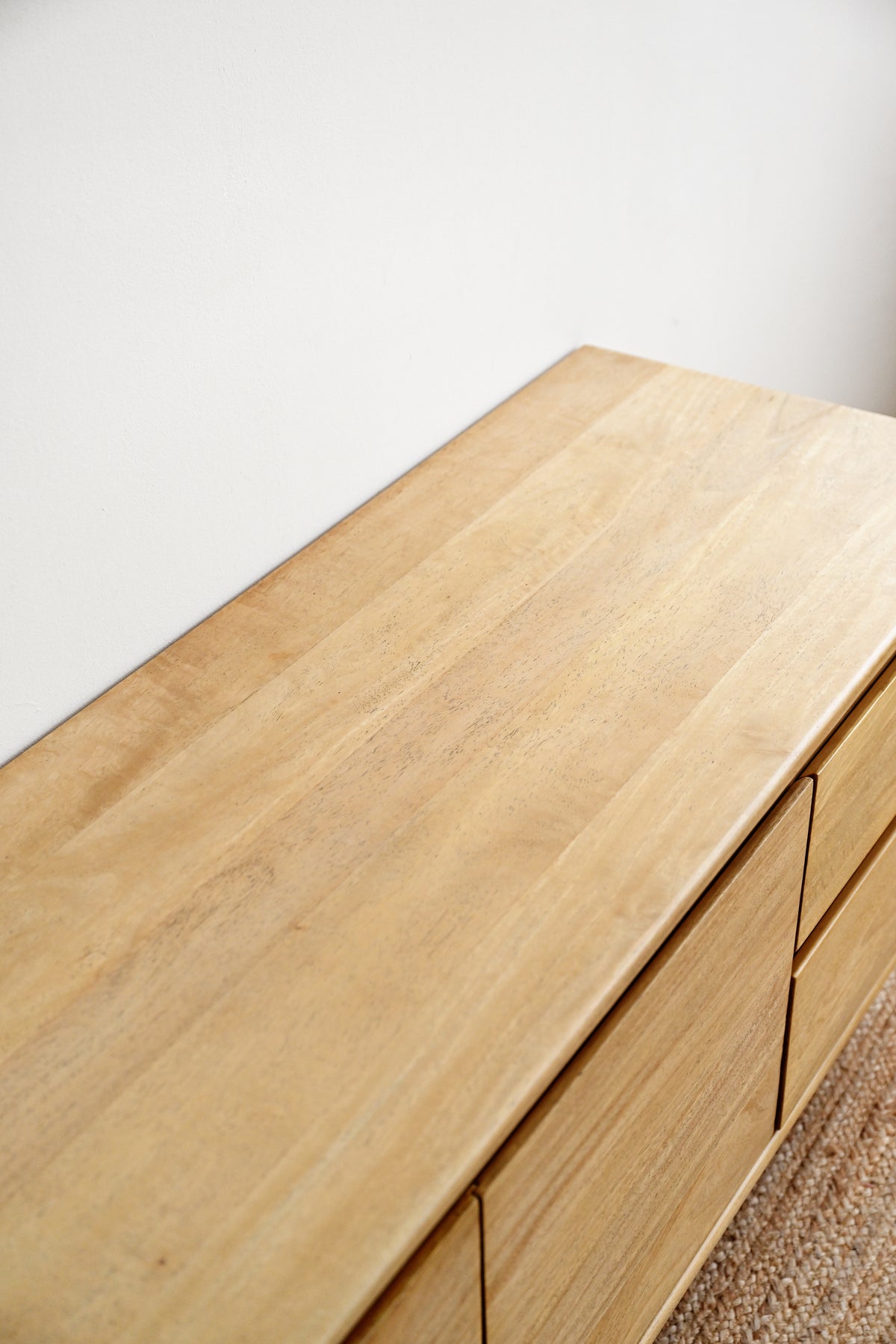 Close-up of a light natural wood dresser or cabinet with drawers, shown against a plain off-white wall and a small section of woven rug.