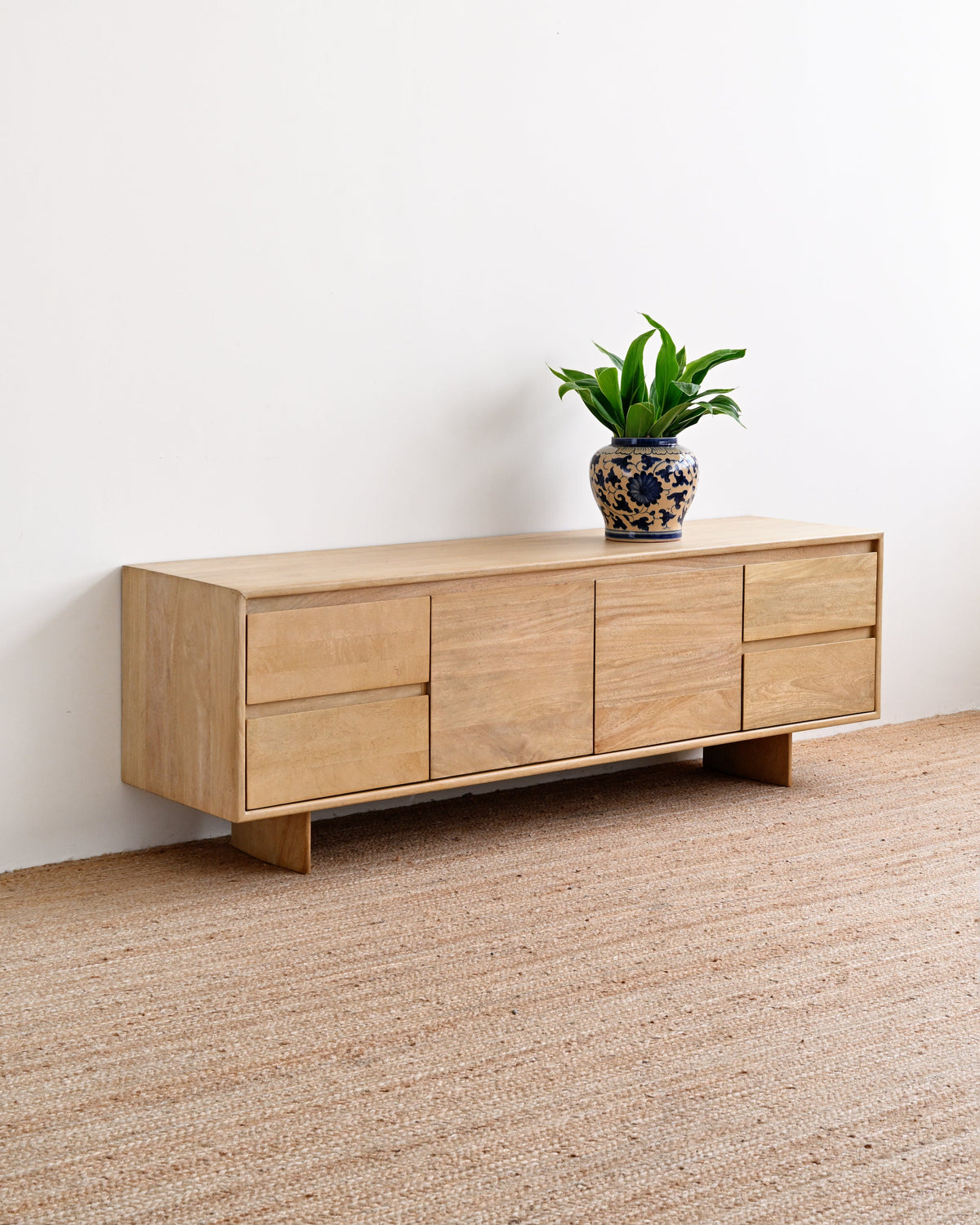 A light natural wood sideboard or credenza with multiple drawer fronts, shown in a minimalist room setting with a woven jute rug and a small potted green plant on top.