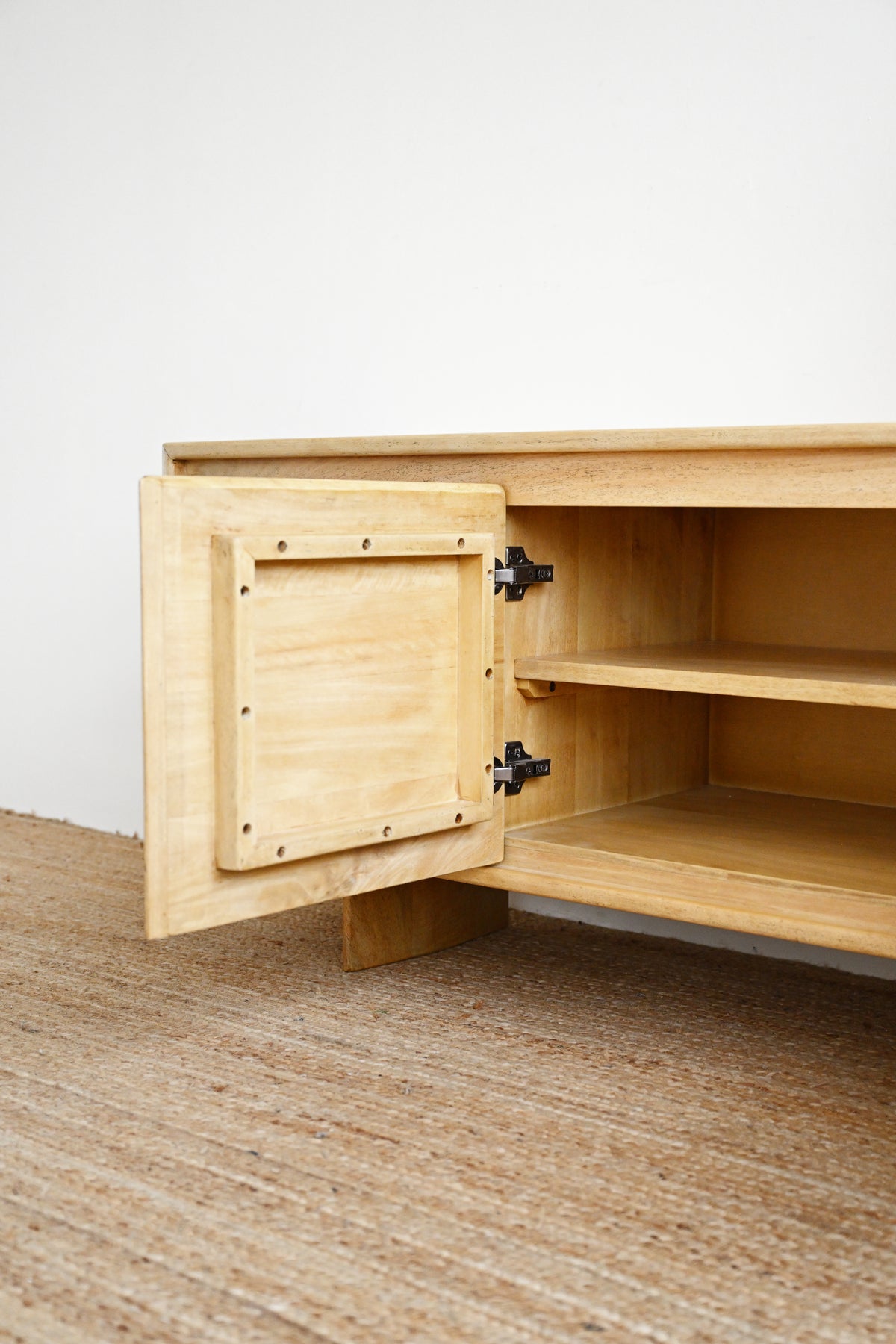 Close-up of a light natural wood cabinet with one door swung open, showing black metal hinges and interior shelves, set against a plain white wall and woven jute flooring.