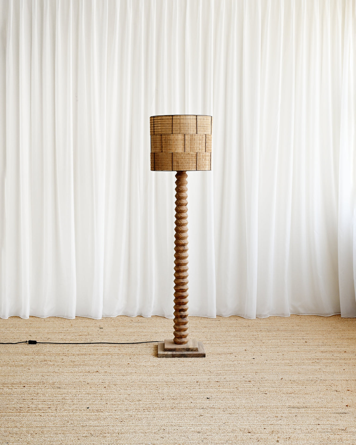 A tall floor lamp with a woven natural-fiber drum shade and a twisted wooden pedestal base, standing on a textured beige rug in front of white draped curtains.