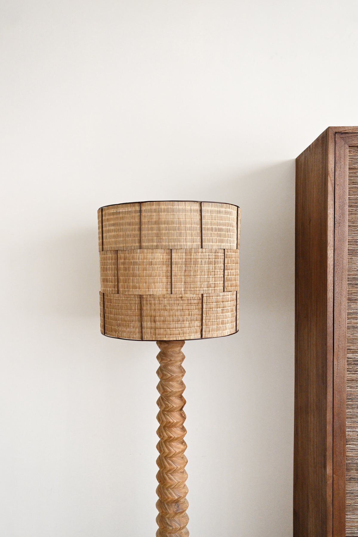 A floor lamp with a cylindrical woven natural-fiber shade and a twisted wooden base, shown against a plain off-white wall with part of a wooden cabinet visible on the right.