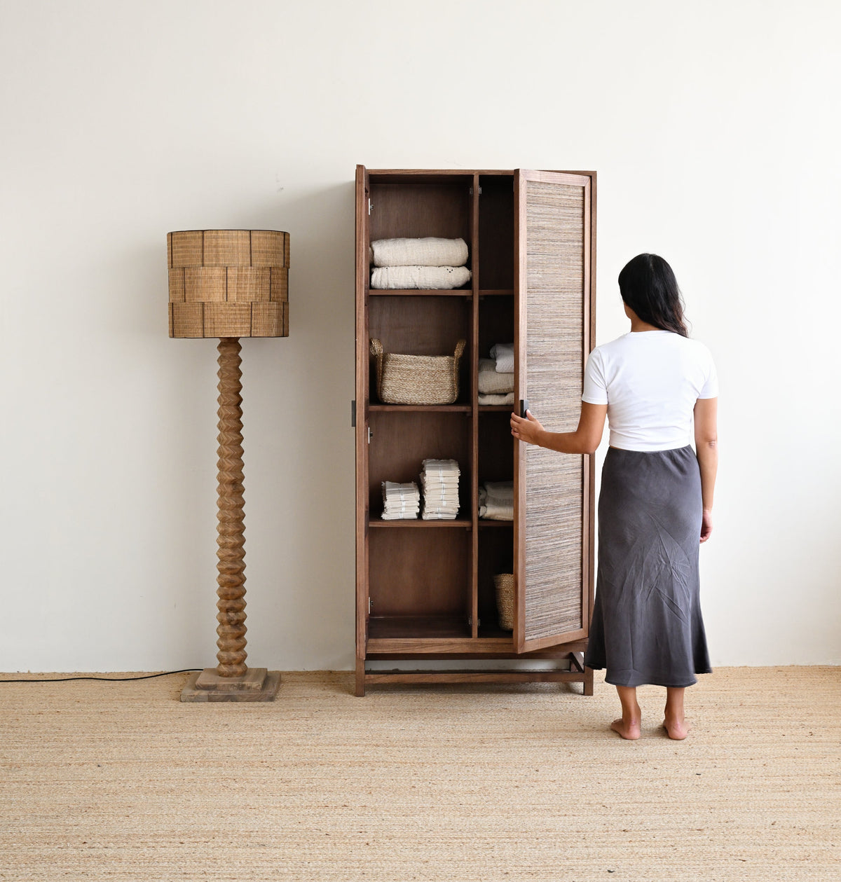 A woman stands in front of a tall wooden cabinet with slatted doors, opening one side to reveal folded white linens inside. The cabinet is placed in a bright room with a woven floor lamp beside it.