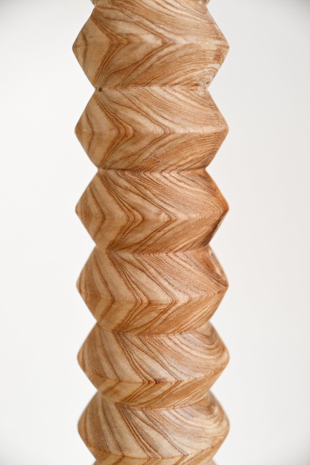 A close-up of a sculptural wooden vase or candle holder with a stacked, geometric, faceted design and a prominent natural wood grain pattern in warm tan and brown tones, set against a plain white background.