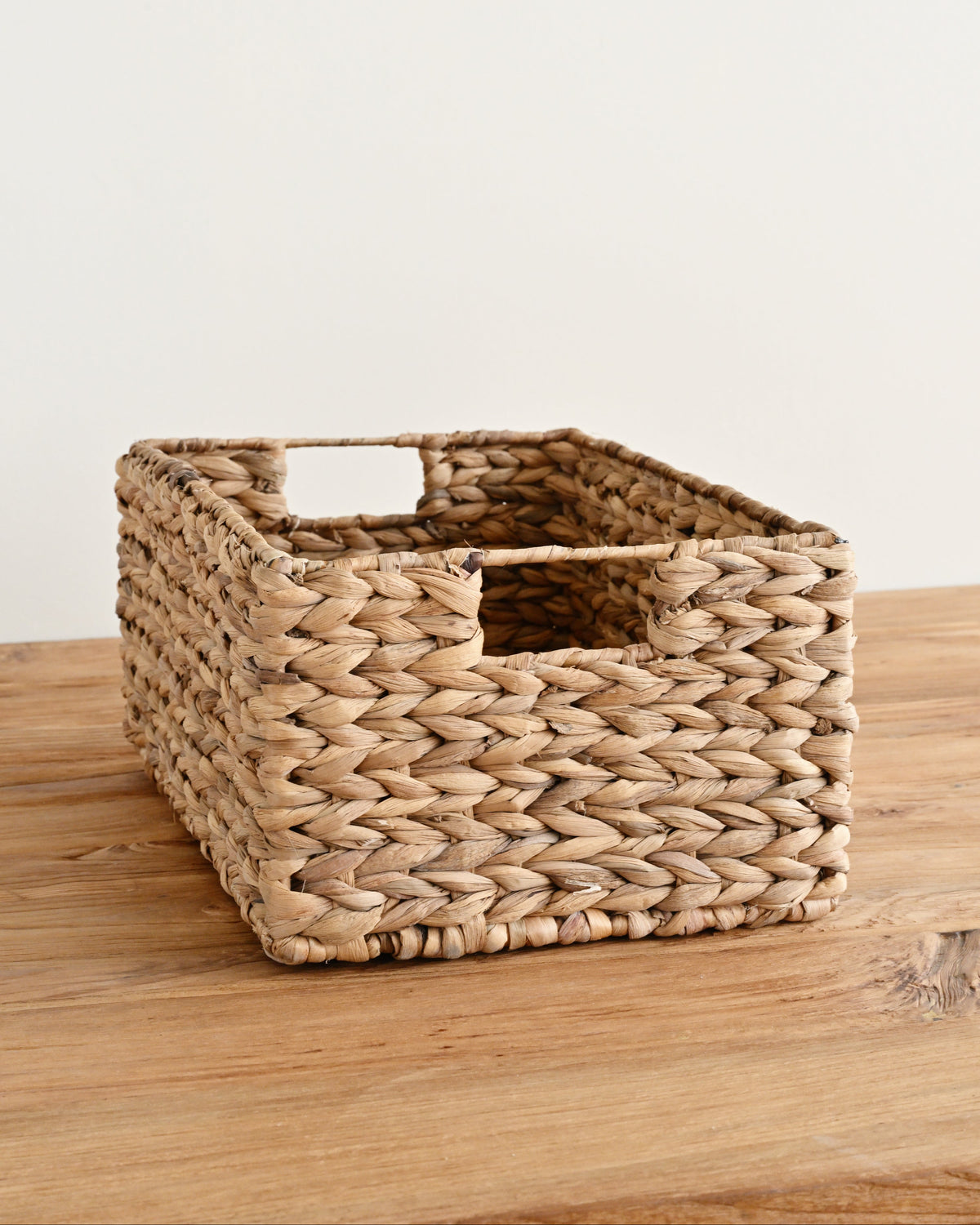 A rectangular woven storage basket made from natural seagrass or wicker, featuring integrated handles, resting on a light wood surface against a plain off-white wall.