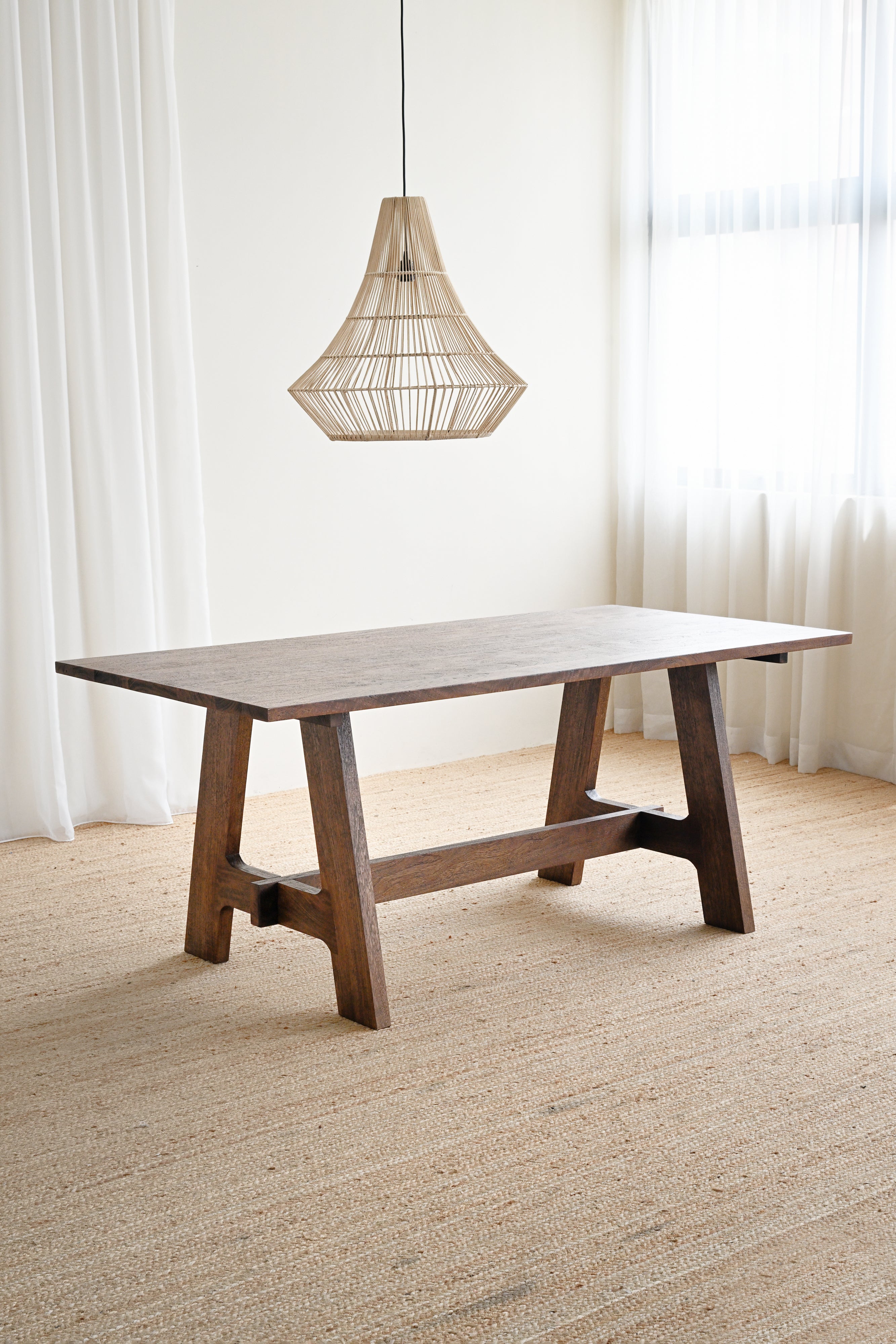 Wooden dining table with a hanging light fixture in a room with white curtains and beige carpet.