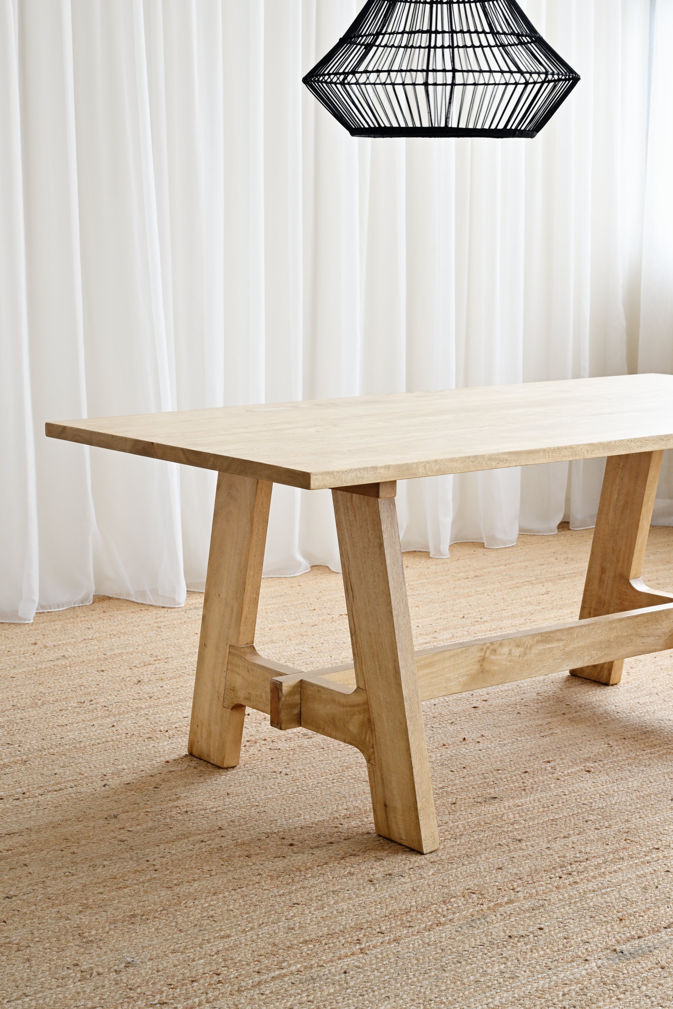 Wooden dining table with a black lampshade in a room with white curtains and a textured floor.