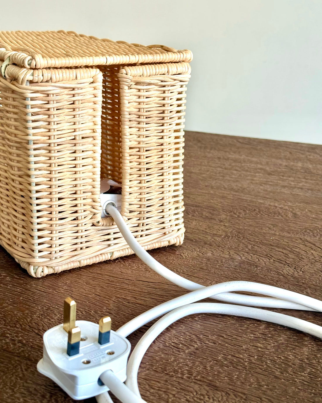 A natural woven rattan or wicker storage basket with a lid sits on a brown wooden table, with a white power cord plugged into it and trailing across the surface.