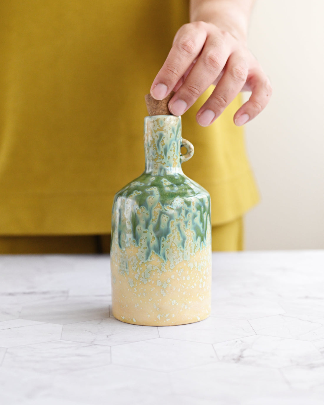 Person holding a ceramic bottle with green and beige design on a white surface.