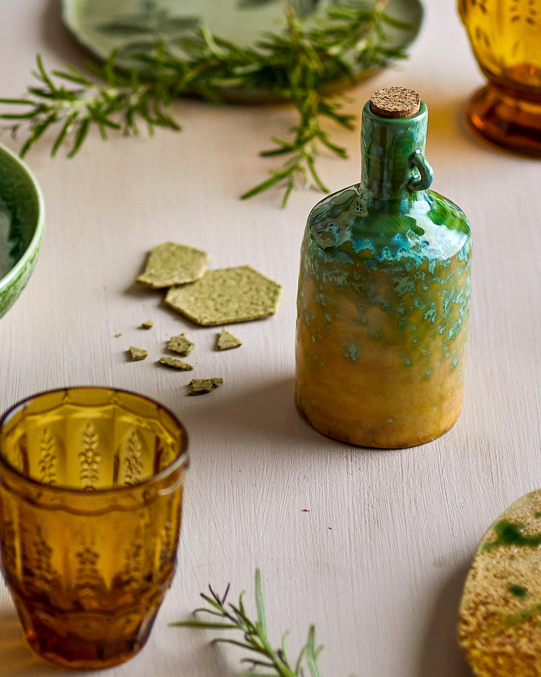 Green ceramic bottle with cork stopper on a light surface with greenery and glasses.