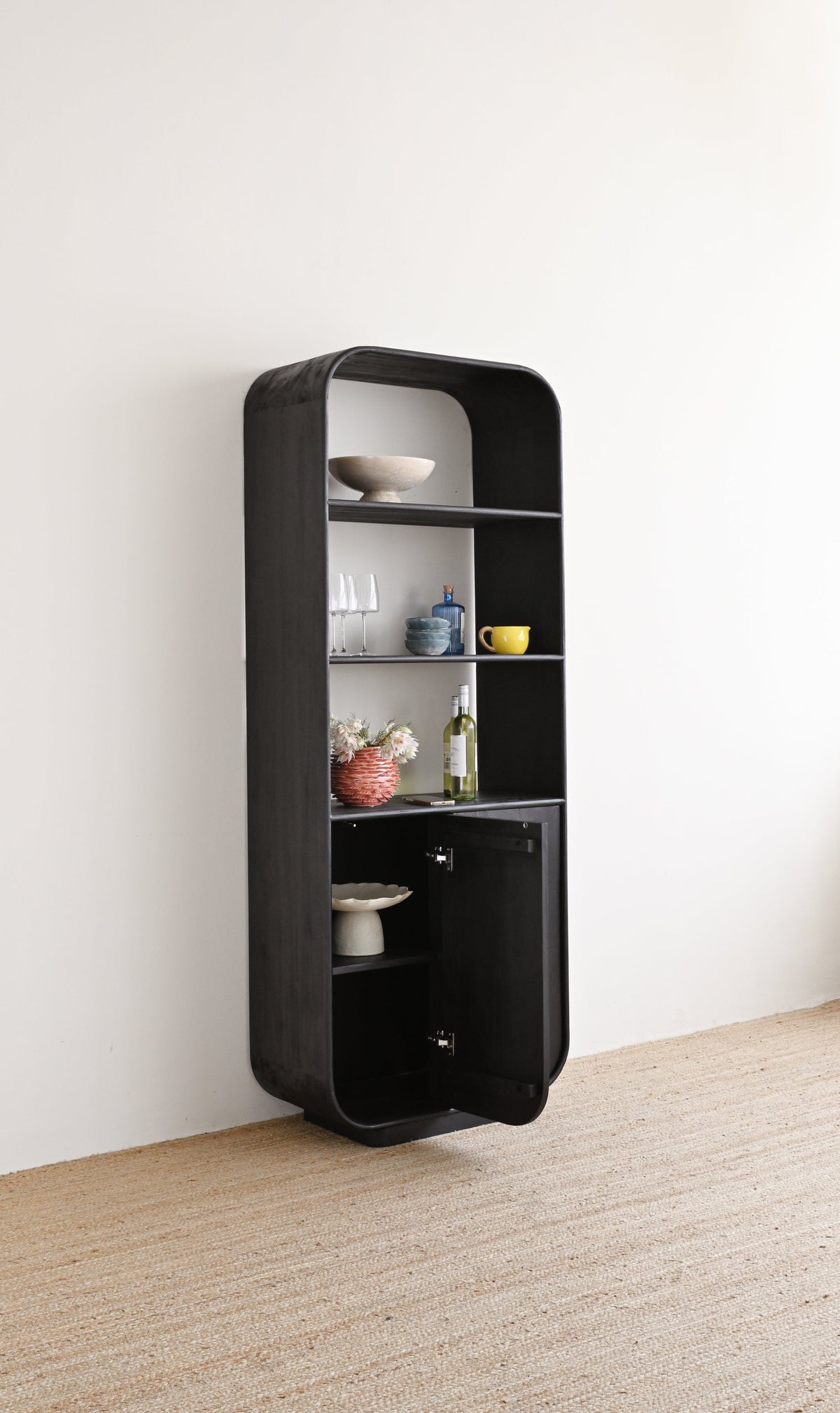 A tall, narrow black shelving unit with rounded edges, open shelves, and a lower cabinet door, styled with small decorative objects. It is photographed in a minimalist room with a white wall and light beige textured floor.