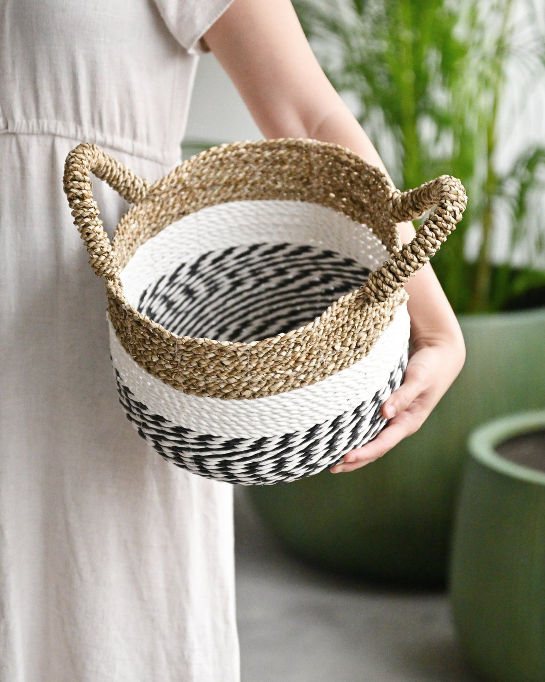A person holding a round seagrass basket with handles, which is predominantly white with black stripes, displayed against a backdrop of indoor plants.
