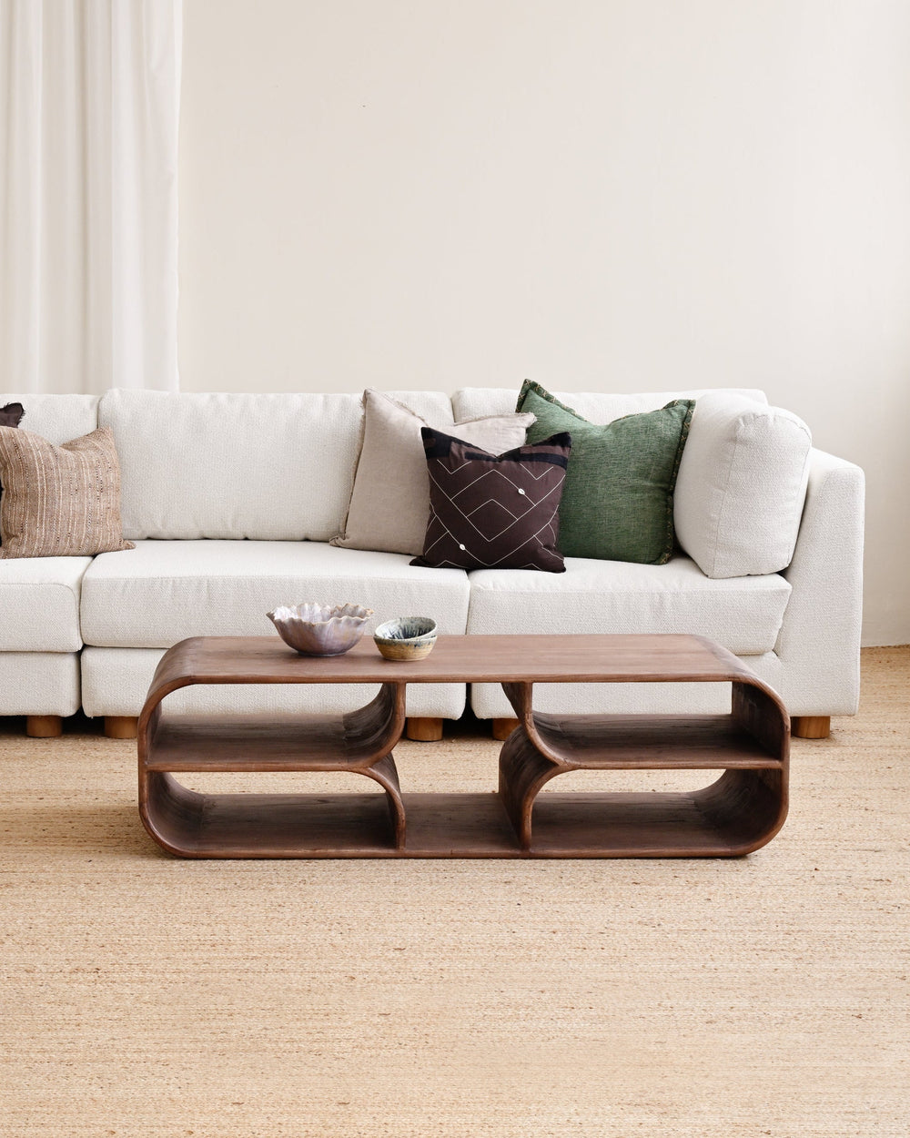 A modern living room setup featuring a white sectional sofa with multiple accent pillows, a matching white ottoman, and a sculptural dark brown wooden coffee table on a light beige jute rug.