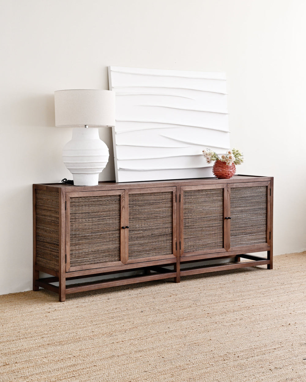 A mid-century modern wooden credenza or sideboard with woven rattan doors, styled in a room setting with a white table lamp, a white textured canvas print, and a small potted plant on top.