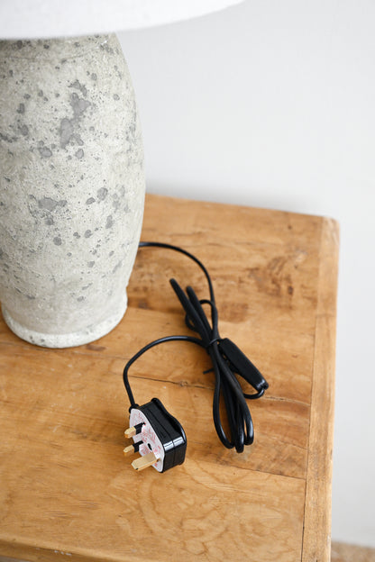 Black power cord with plug on a wooden surface next to a textured white vase.