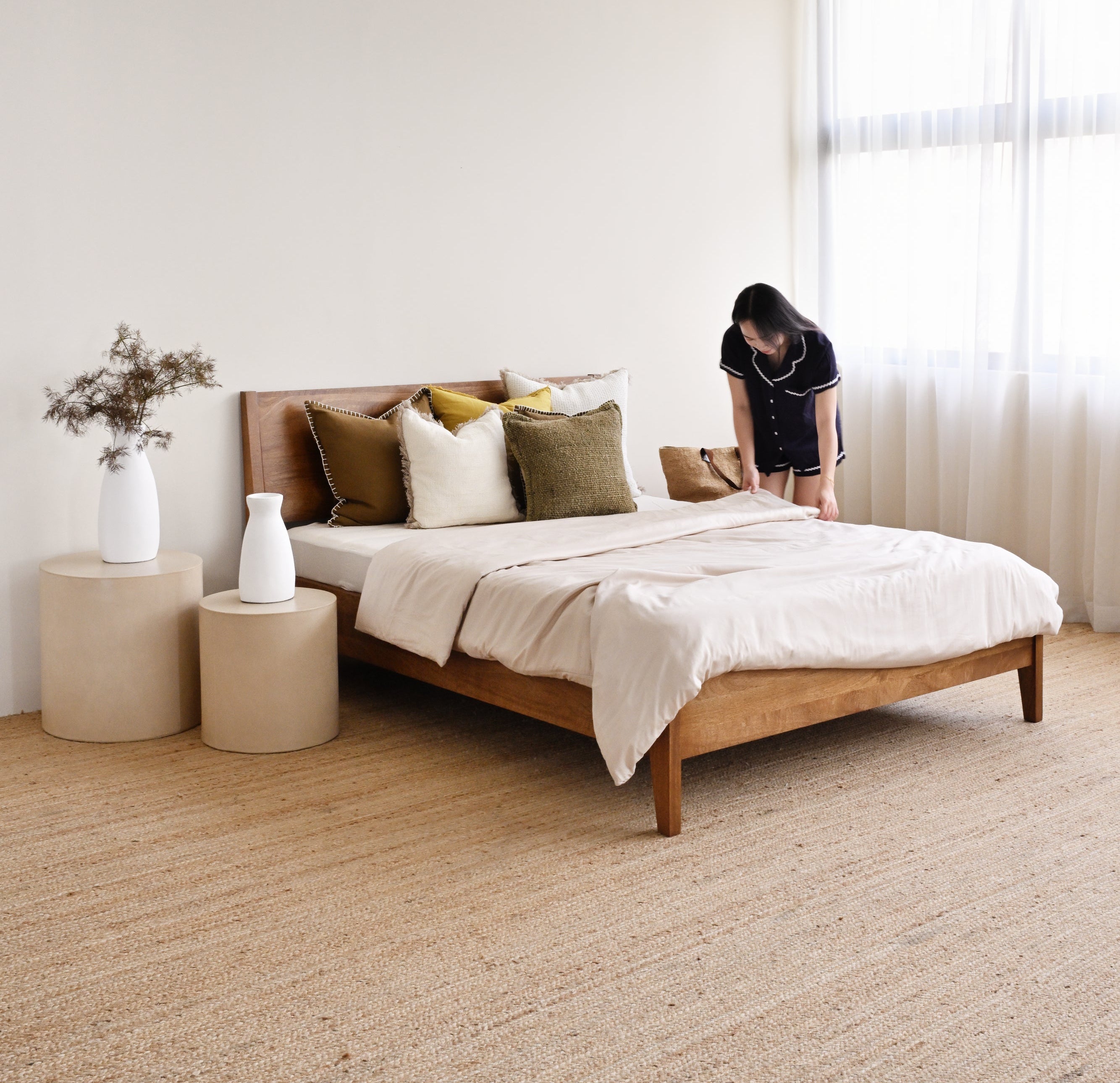 Person standing next to a wooden bed with white bedding and decorative pillows in a bright room.