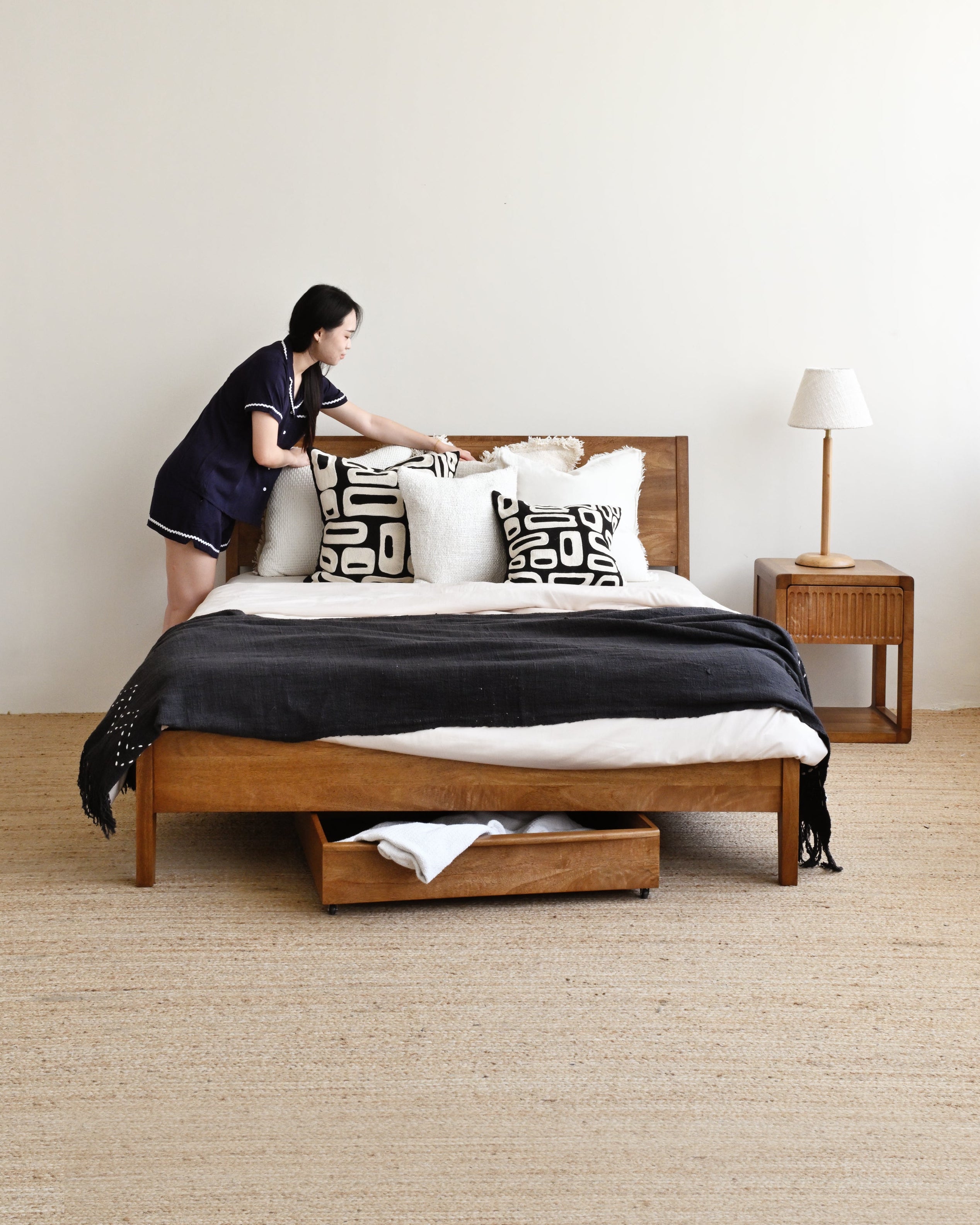 Person making a bed in a modern bedroom with wooden furniture and neutral decor.