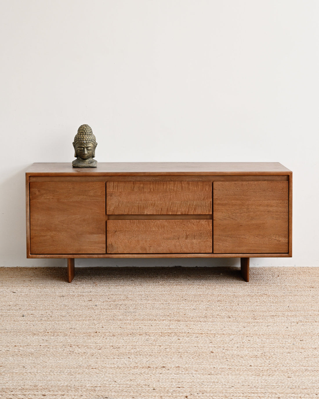 A mid-century modern wooden sideboard with three drawers and two cabinet doors, styled on a textured beige jute rug against a plain white wall with a small Buddha head sculpture on top.