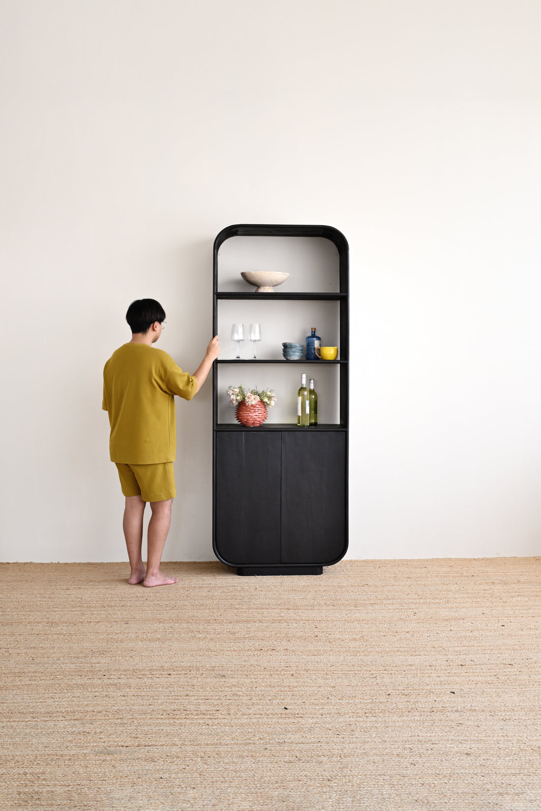 A person in a yellow-green shirt and shorts stands in front of a tall black shelving unit in a minimalist room with a white wall and light beige floor.