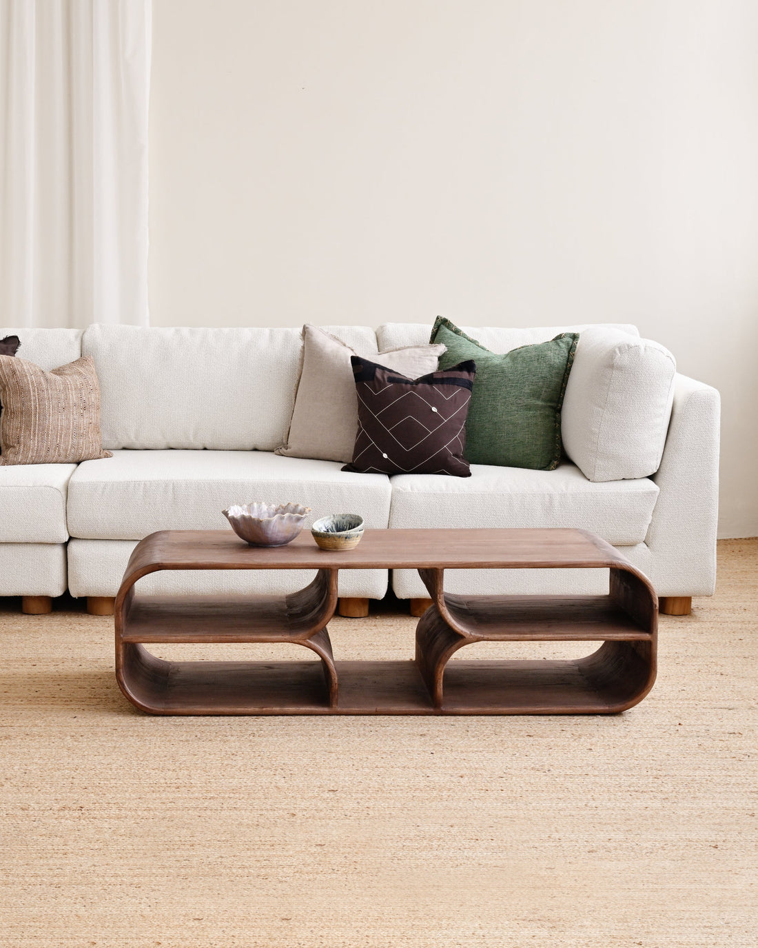 A modern living room setup featuring a white sectional sofa with multiple accent pillows, a matching white ottoman, and a sculptural dark brown wooden coffee table on a light beige jute rug.
