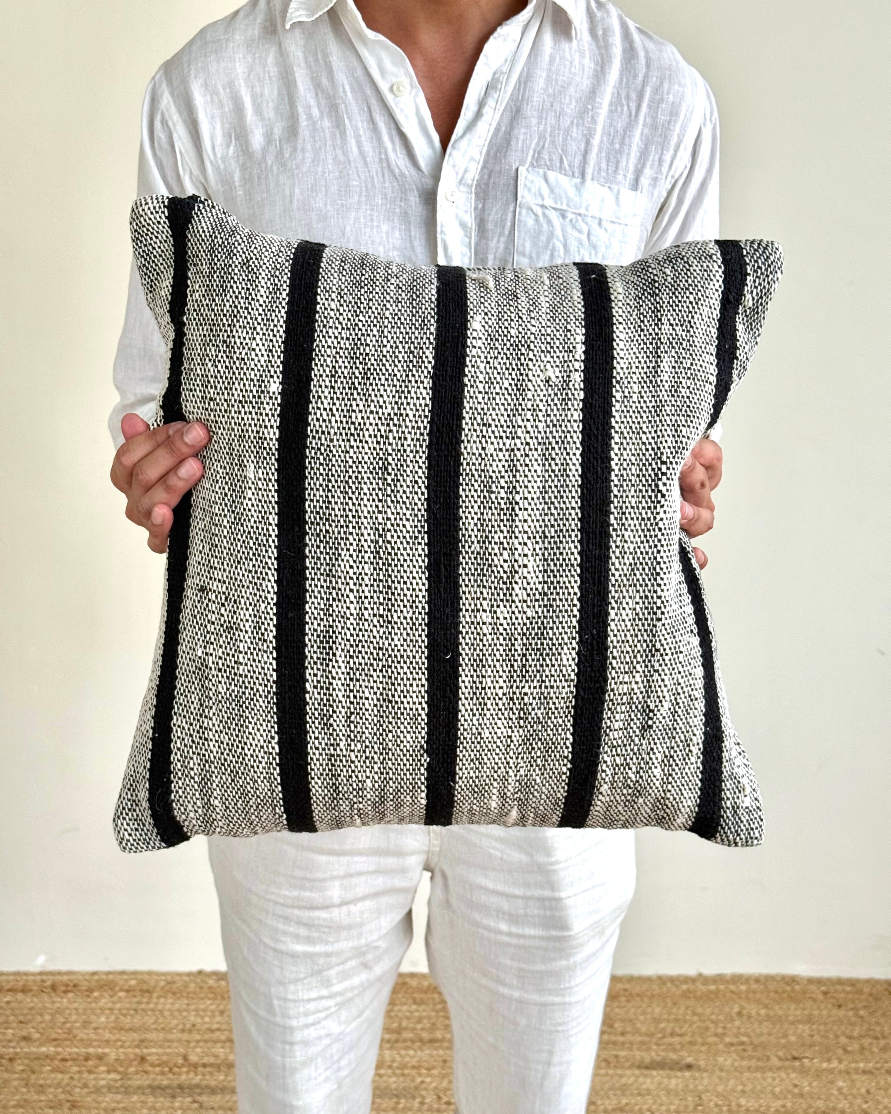 A person wearing a white linen shirt and white pants holds a square woven throw pillow with vertical black stripes on a textured gray and white fabric background.
