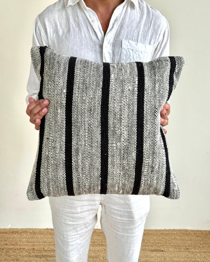 A person wearing a white linen shirt and white pants holds a square woven throw pillow with vertical black stripes on a textured gray and white fabric background.