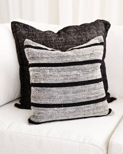 A square throw pillow with horizontal black stripes on a textured gray and white woven fabric, styled on a white sofa in front of a dark charcoal pillow.