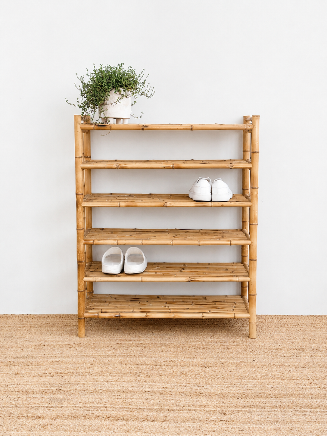 Bamboo shoe rack with shoes and a plant on a wooden floor against a white wall.