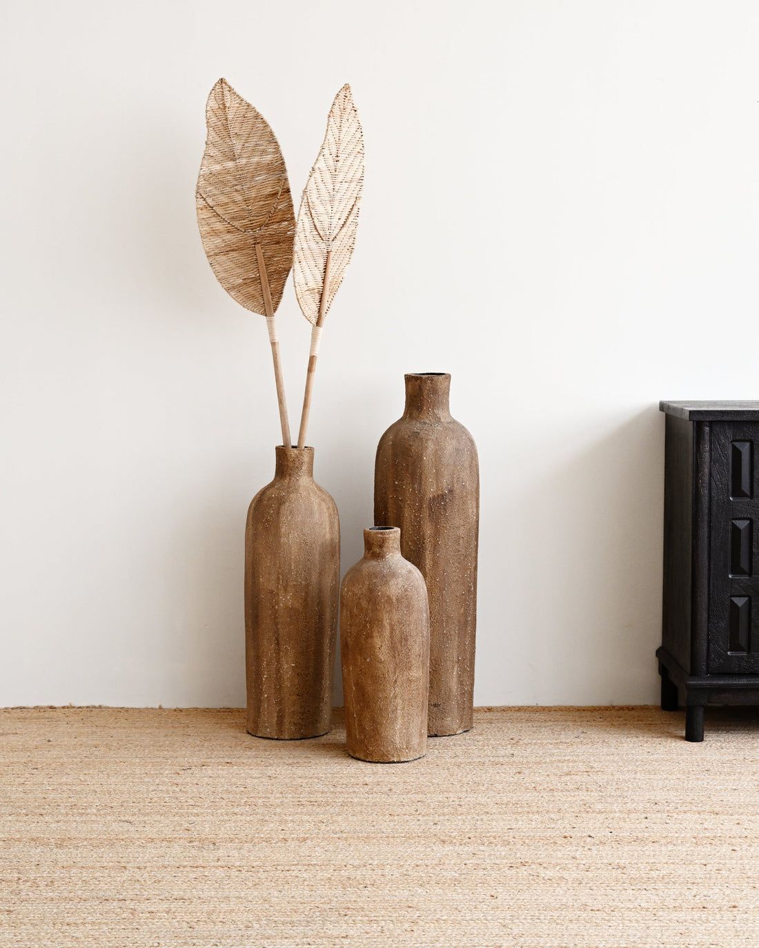 Three wooden vases with dried leaves on a beige carpet against a white wall.