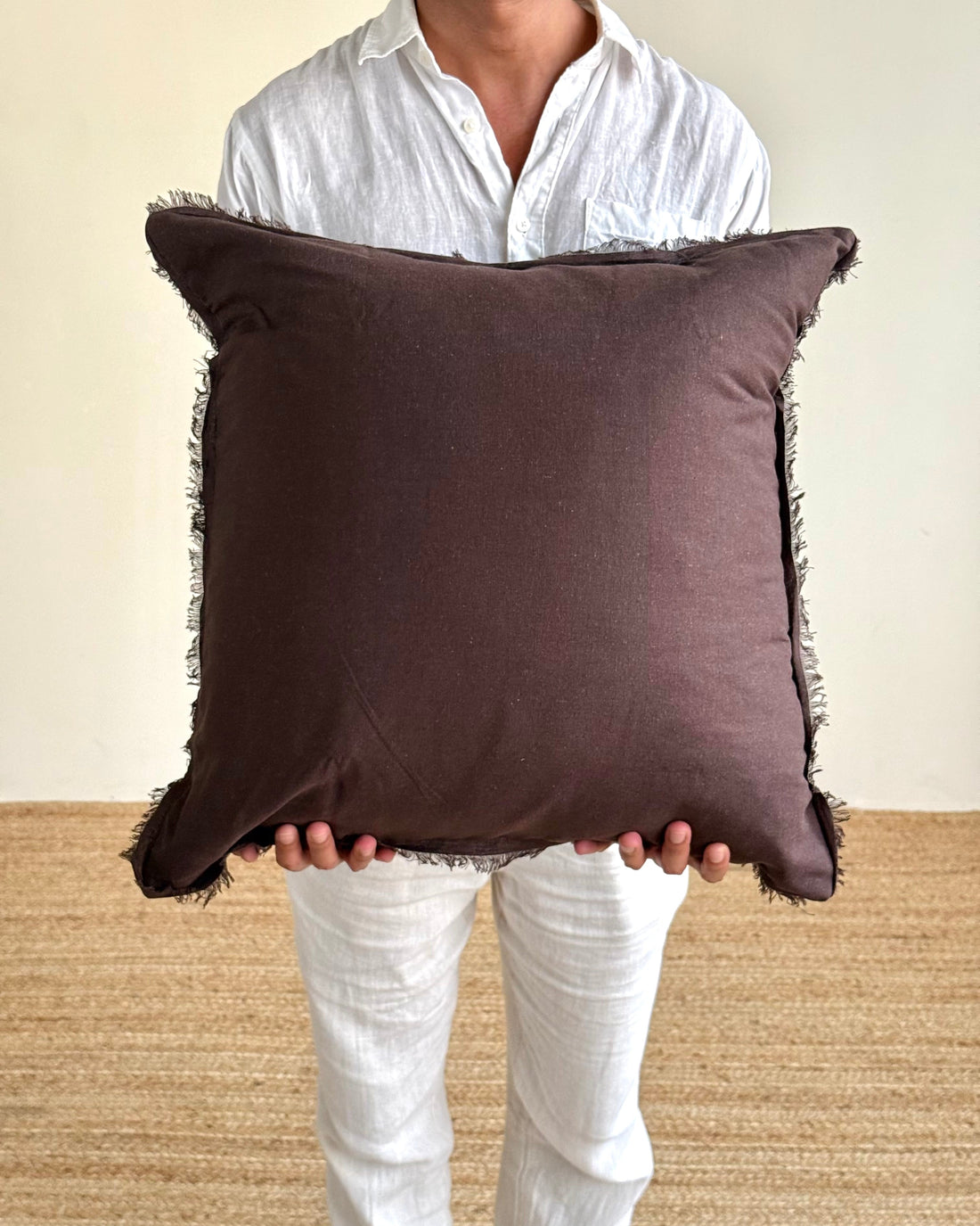 A person wearing a white shirt and white pants holds a square dark brown throw pillow with fringed edges against a plain beige wall and woven floor.