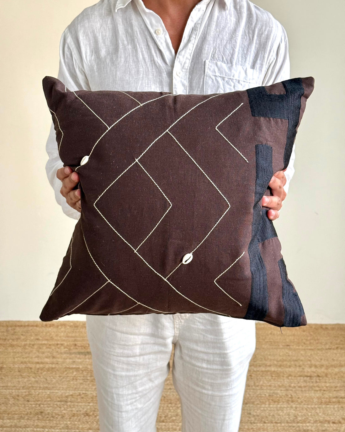 A person in a white shirt and white pants holds a square brown throw pillow with a white geometric line pattern and a dark blue abstract patch on the right side, against a neutral beige background.
