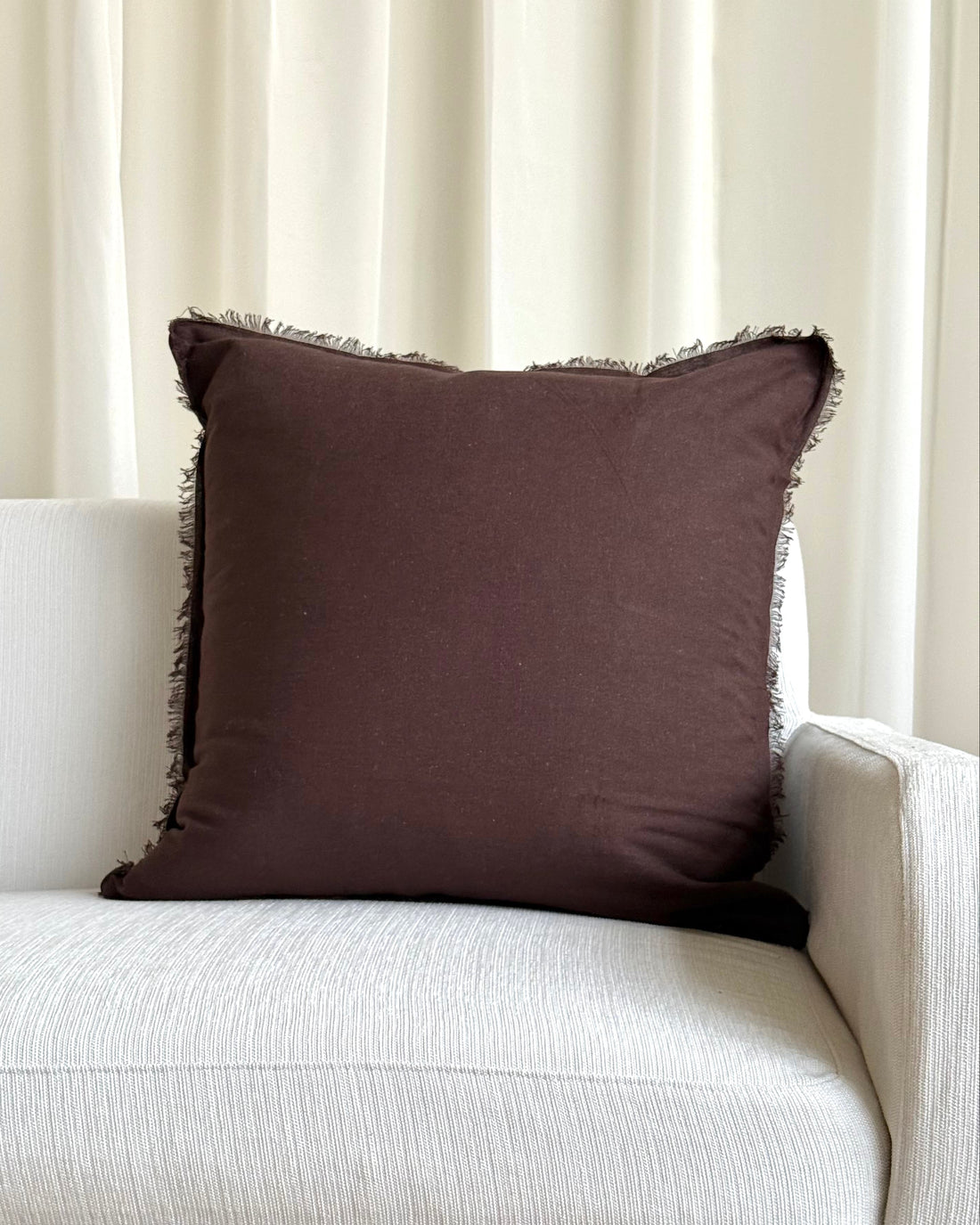 A square dark brown throw pillow with fringed edges resting on a light beige textured sofa in front of cream-colored curtains.