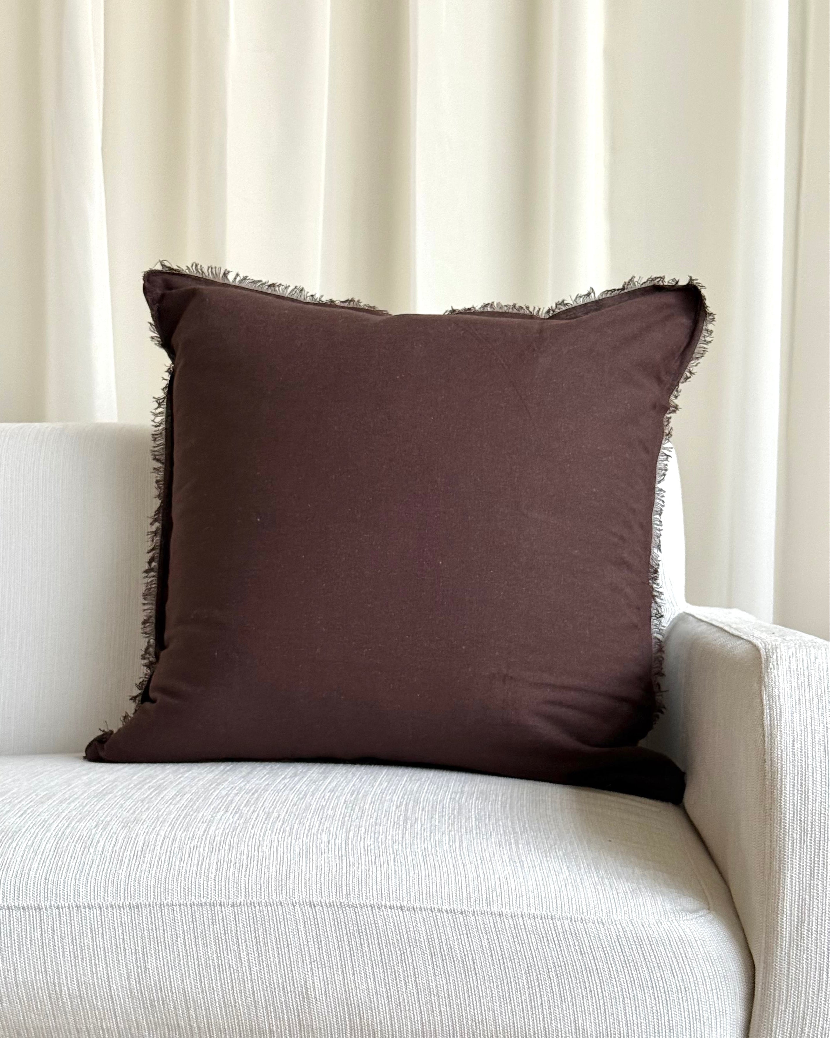 A square dark brown throw pillow with fringed edges resting on a light beige textured sofa in front of cream-colored curtains.