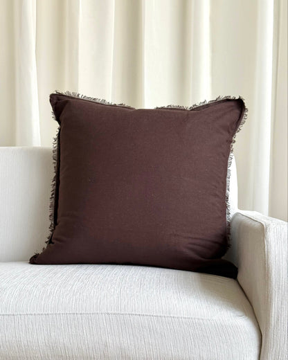 A square dark brown throw pillow with fringed edges resting on a light beige textured sofa in front of cream-colored curtains.