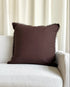 A square dark brown throw pillow with fringed edges resting on a light beige textured sofa in front of cream-colored curtains.