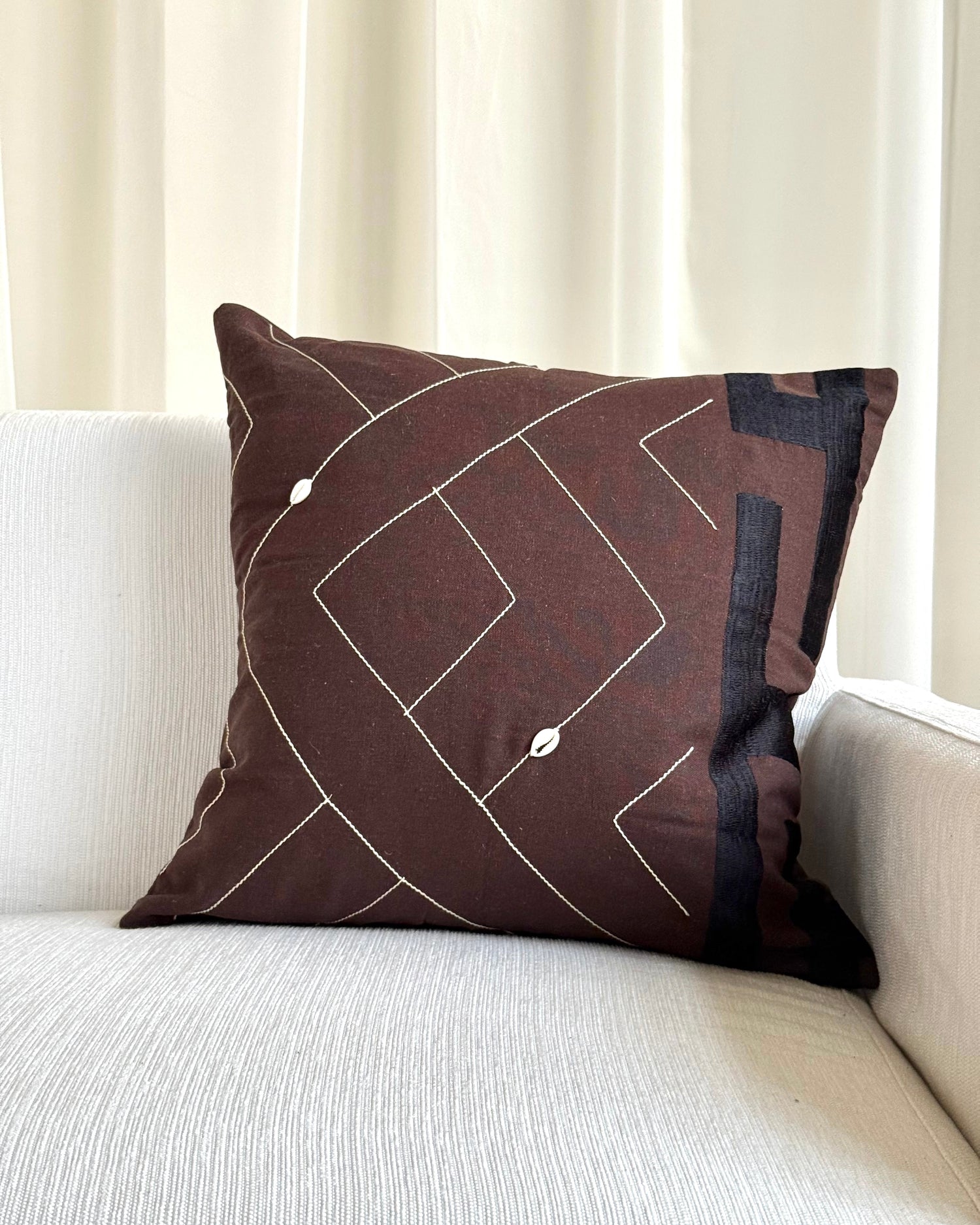 A square brown throw pillow with a white geometric line pattern and small white bead accents, resting on a light beige textured sofa in front of cream-colored curtains.