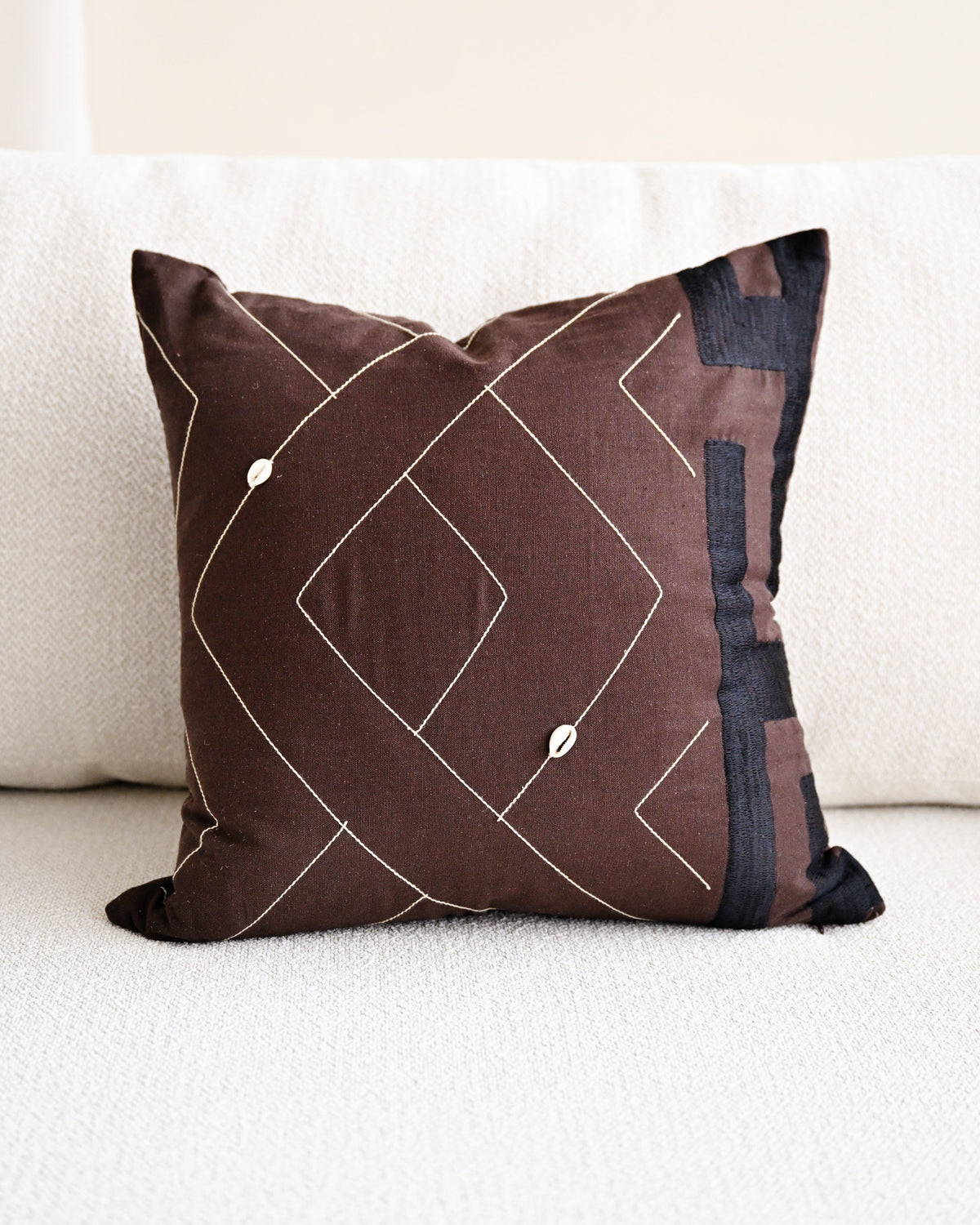 A square brown throw pillow with a geometric diamond pattern in white and dark navy blue accents, resting on a white textured sofa.