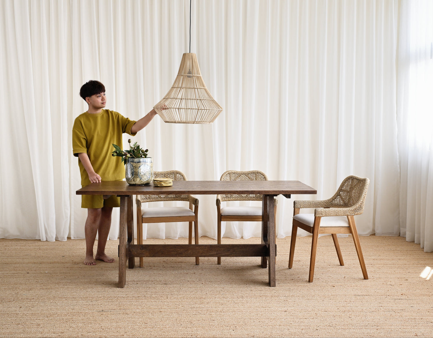 Person adjusting a pendant light over a wooden dining table with chairs in a room with white curtains.